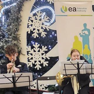 Three young people playing brass instruments, sitting in front of Christmas decorations.